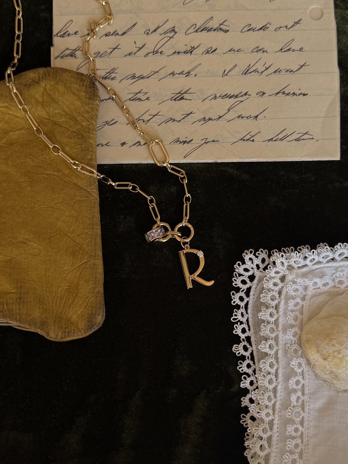 The Mod + Jo Este Birthstone Charm Spacer, a personalized 18k gold plated necklace with an R charm, rests on handwritten letters, a gold pouch, white lace-trimmed handkerchief, and a stone on dark fabric.