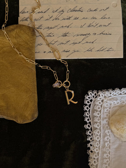 The Mod + Jo Este Birthstone Charm Spacer, a personalized 18k gold plated necklace with an R charm, rests on handwritten letters, a gold pouch, white lace-trimmed handkerchief, and a stone on dark fabric.