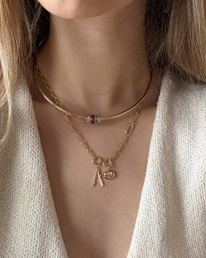 A woman wears two gold Mod + Jo necklaces: a choker with three colorful gemstones and a chain with an “A” pendant, a small round charm, and the Este Birthstone Charm Spacer. She pairs them with a cream textured top.