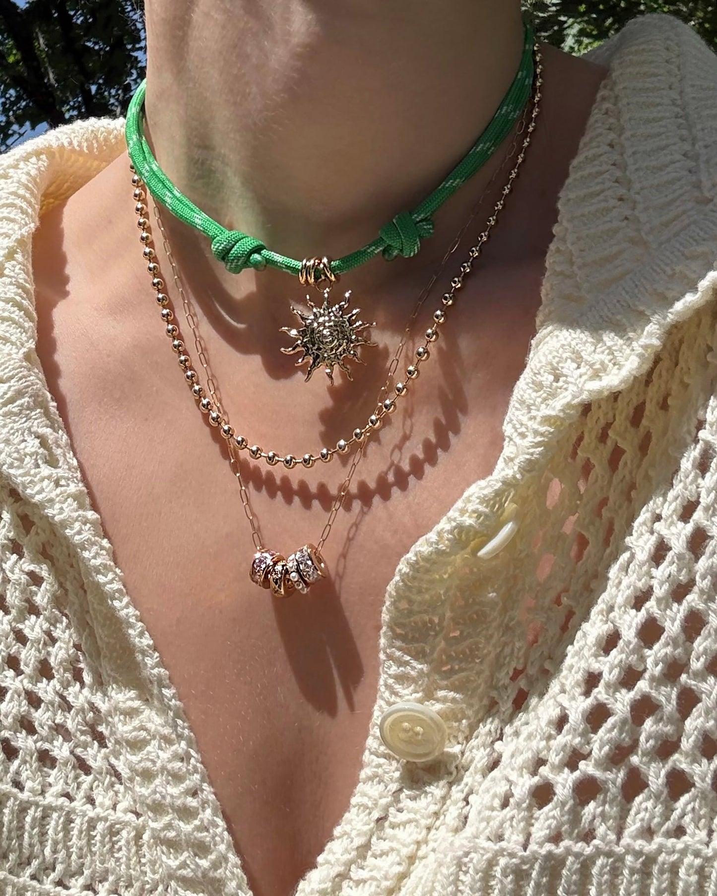 A person wears a cream crochet top and three layered necklaces: the Mod + Jo Tommy Cord Necklace, plus a gold chain with small beads, and a delicate chain with three silver and gold bead charms.
