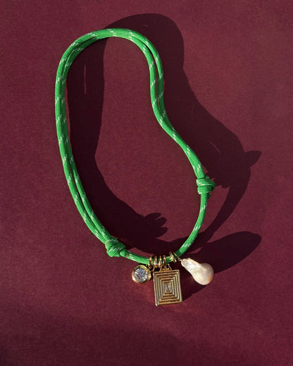 The Mod + Jo Tommy Cord Necklace features a green paracord and three charms—a round gem, a geometric gold pendant, and a white pearl—shown on a burgundy background.