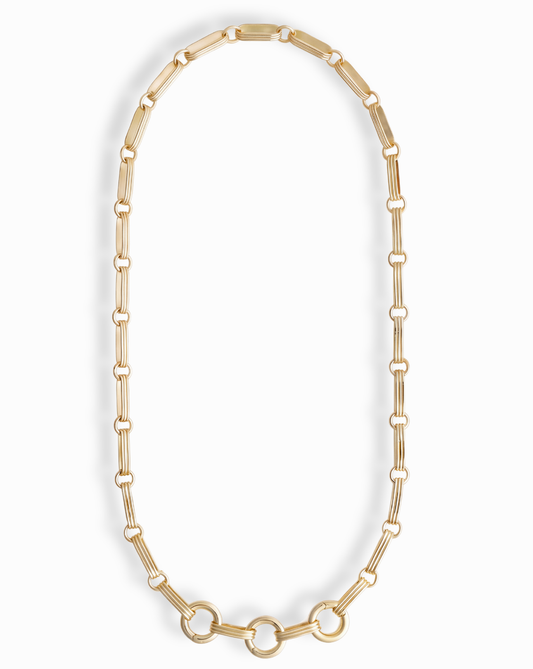 The Jagger Charm Clip Necklace by Mod + Jo features elongated oval links crafted in 14k gold and showcases three larger connected circles at the bottom, elegantly displayed on a white background. This sophisticated necklace seamlessly blends timeless style with contemporary flair.