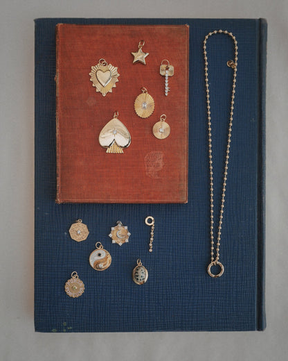 The Soren Ball Charm Clip Necklace by Mod + Jo is displayed on two stacked books, one red and one blue, adorned with gold charms in various shapes such as a heart, star, key, sun, yin-yang, and more—all arranged neatly for viewing.