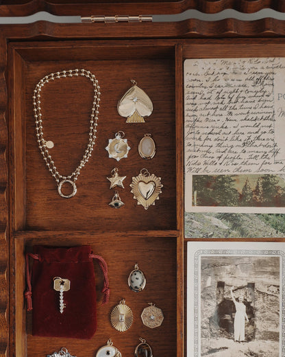 A wooden jewelry box contains gold charms, a bracelet, a protective Mod + Jo Nyx Evil Eye Charm pendant, a red velvet pouch holding a key, vintage postcards, and a black-and-white photograph of a person standing outside against a stone wall.