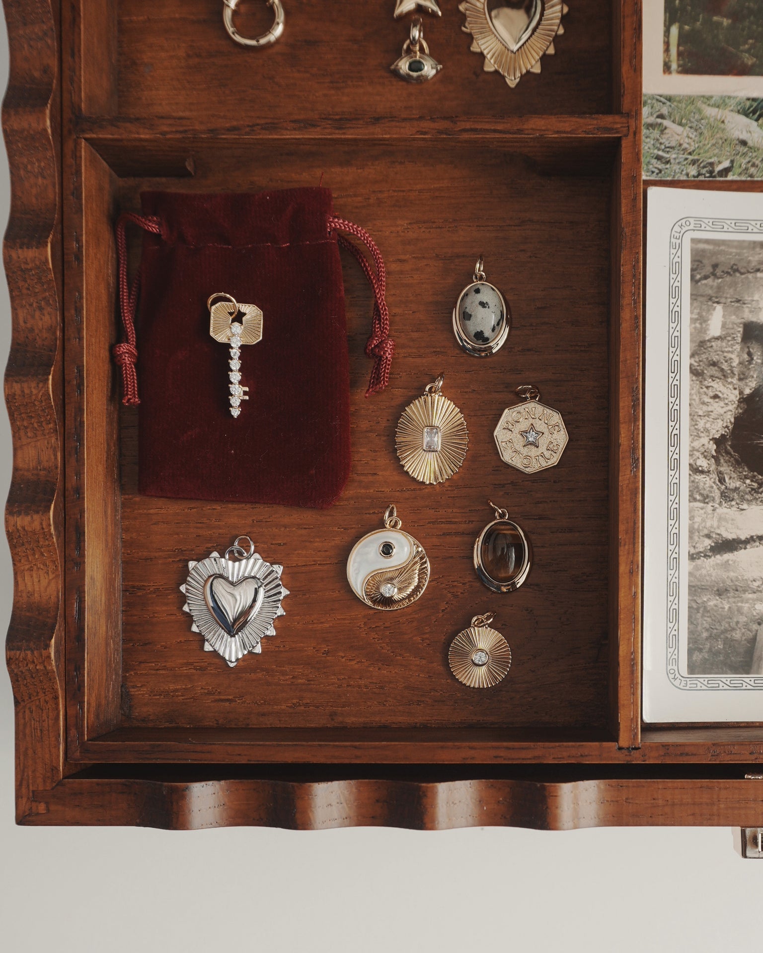 A wooden drawer holds an assortment of vintage charms and pendants, including the Grande Ava Yin Yang Charm from Mod + Jo, nestled among heart-shaped, spiral, and sunburst designs. A small red velvet pouch and a glimmering mother-of-pearl necklace rest beside the trinkets, while black-and-white photos peek in from the right.