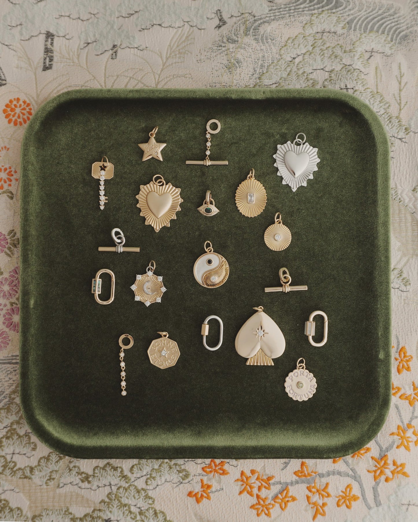 Nora Lock Carabiner charms by Mod + Jo—crafted in gold and silver finishes, featuring a range of shapes like hearts, stars, a yin-yang motif, and geometric forms (some embellished with natural semi-precious stones such as blue lace agate and highlighted with elegant 14k plating)—are artfully arranged on a dark green velvet tray. The backdrop is a richly patterned floral fabric.