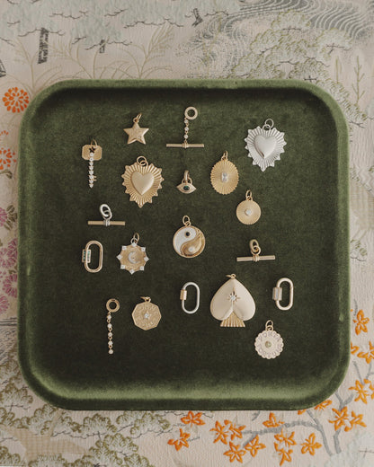 A Nora Lock Carabiner by Mod + Jo is displayed on a green velvet tray alongside 18 assorted gold and silver charms with 14k plating—featuring hearts, stars, circles, a fan, yin-yang, and geometric designs—all arranged on a floral-patterned fabric background.