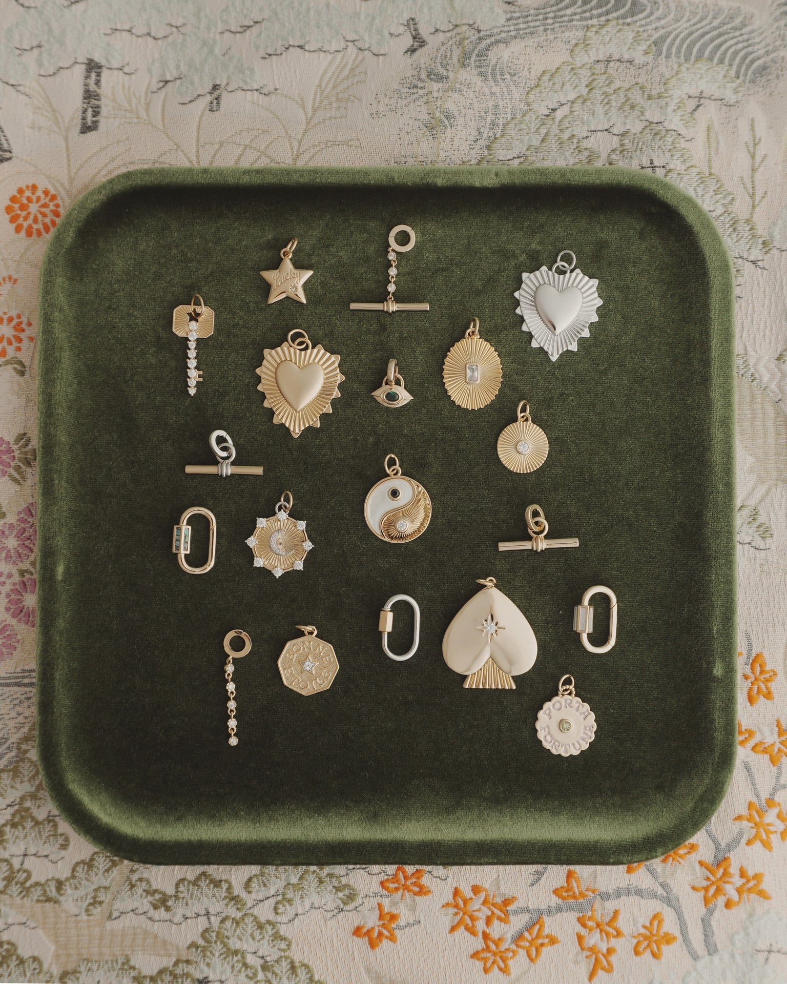 Nora Lock Carabiner charms by Mod + Jo—crafted in gold and silver finishes, featuring a range of shapes like hearts, stars, a yin-yang motif, and geometric forms (some embellished with natural semi-precious stones such as blue lace agate and highlighted with elegant 14k plating)—are artfully arranged on a dark green velvet tray. The backdrop is a richly patterned floral fabric.