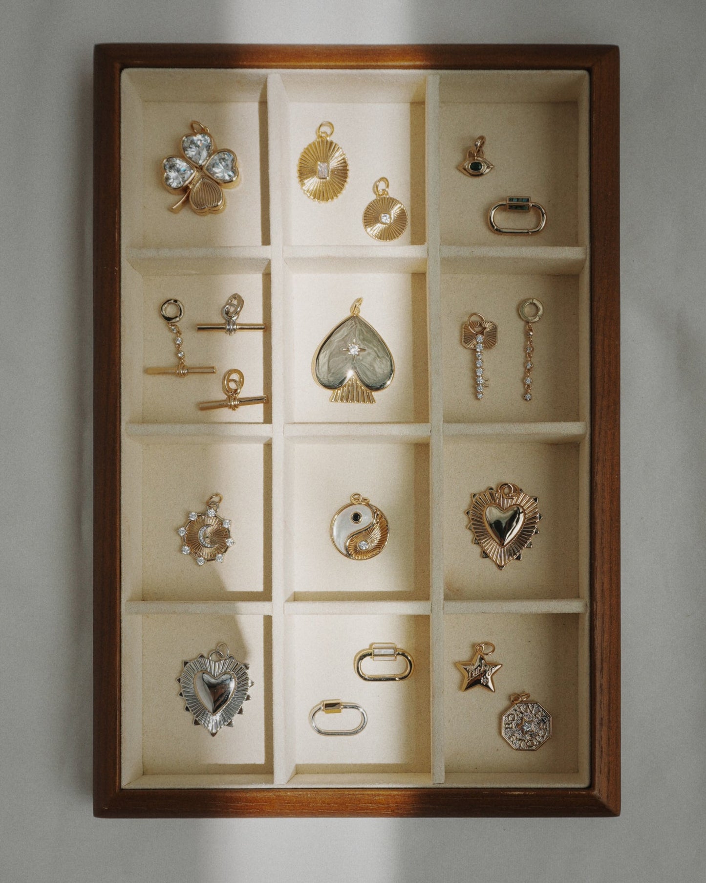 The Mod + Jo Cora Fluted Charm is displayed in a wooden jewelry box with twelve compartments, alongside gold and silver charms such as hearts, stars, an eye, a yin-yang symbol, and various geometric shapes—some highlighted with emerald-cut crystals—all arranged neatly on a cream fabric lining.