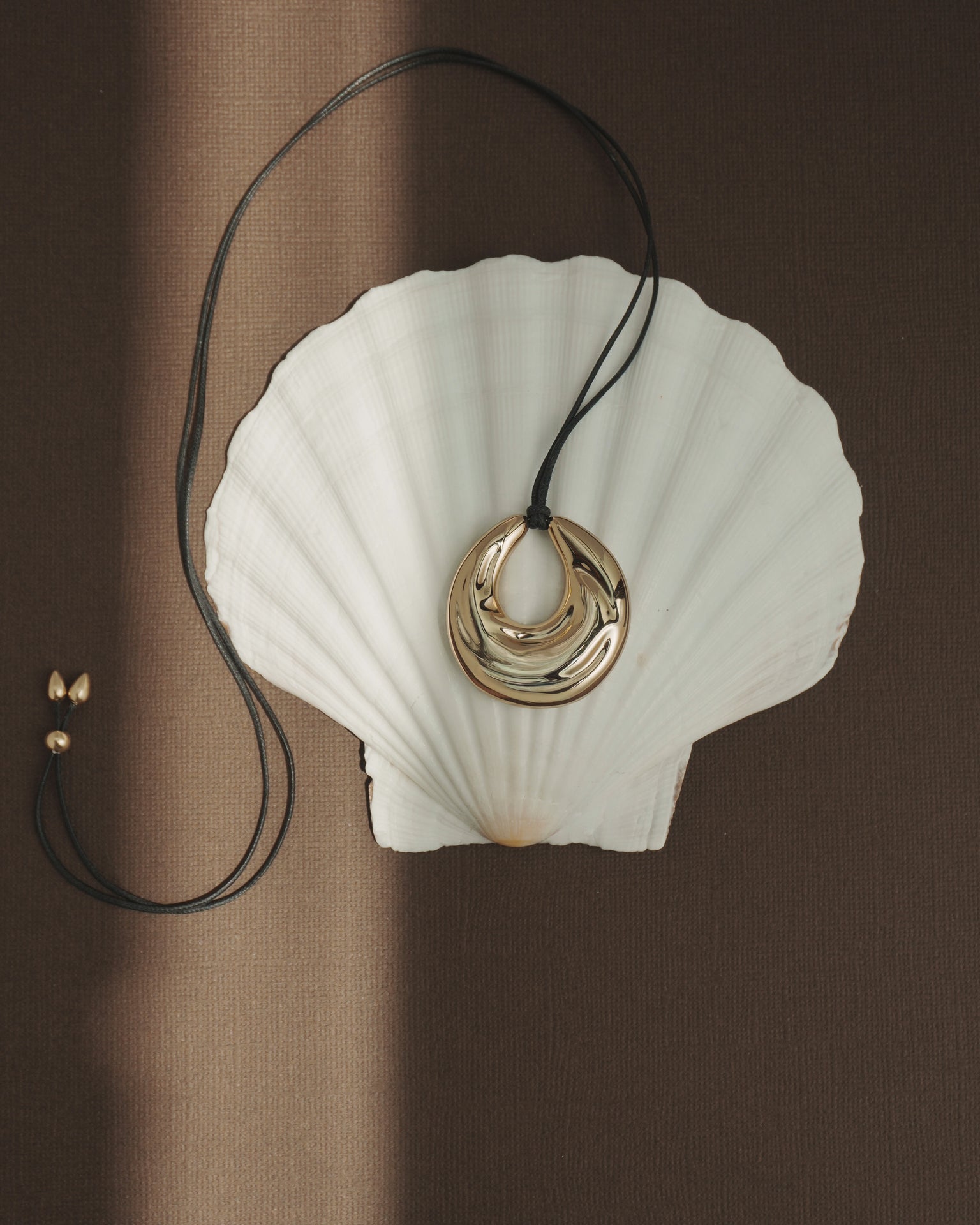 The Kira Cord Necklace by Mod + Jo, featuring a gold pendant on an adjustable cord, is displayed on a white seashell atop a brown textured background with sunlight casting soft shadows across the scene.