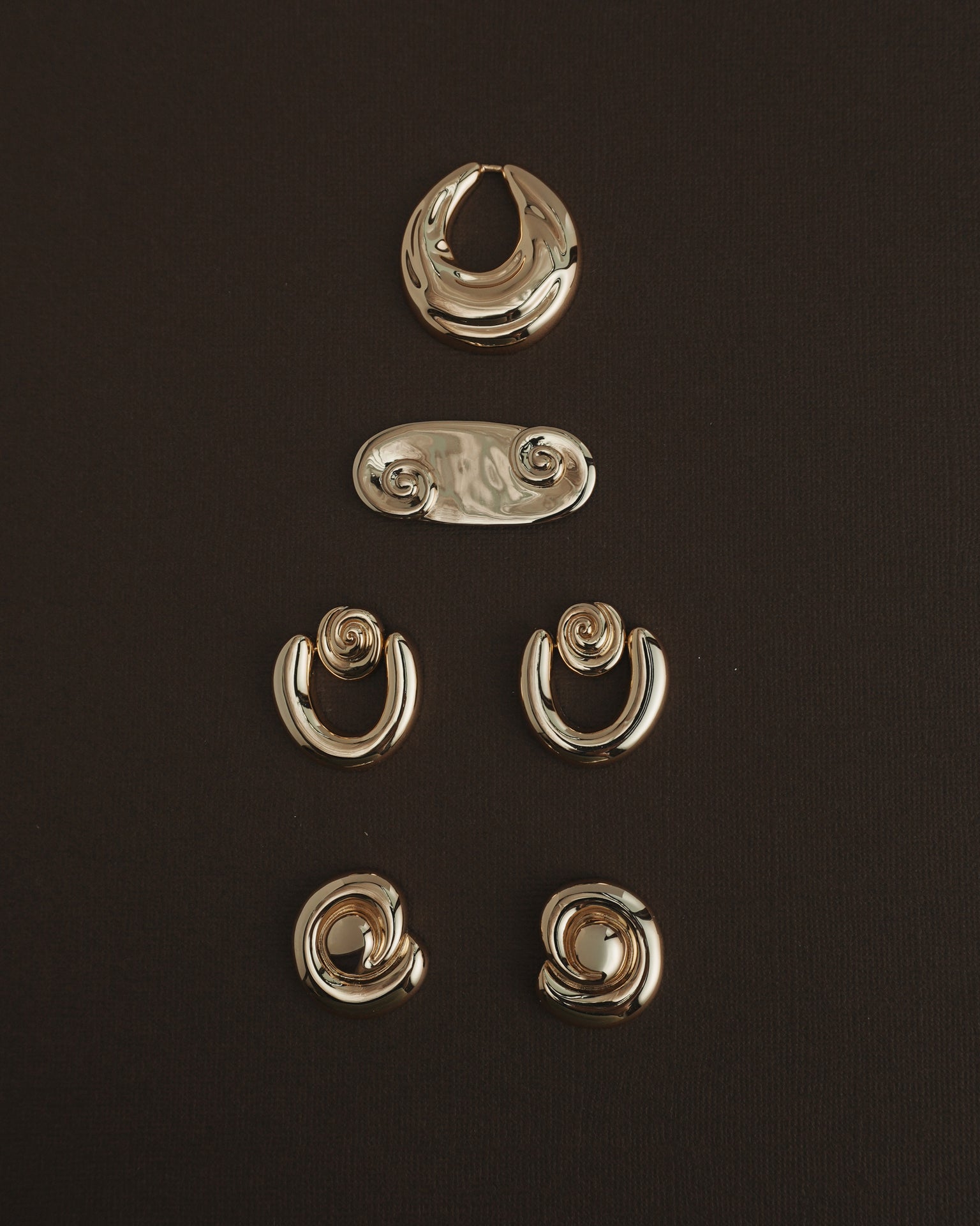Five pairs of Mod + Jo Milley Spiral Statement Studs in 14k gold are artfully arranged on a dark brown background—ideal for daily wear or to add a unique, abstract touch with their swirling design.