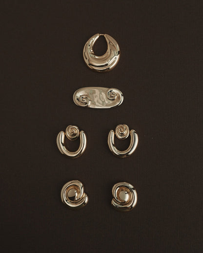 Five pairs of Mod + Jo Milley Spiral Statement Studs in 14k gold are artfully arranged on a dark brown background—ideal for daily wear or to add a unique, abstract touch with their swirling design.
