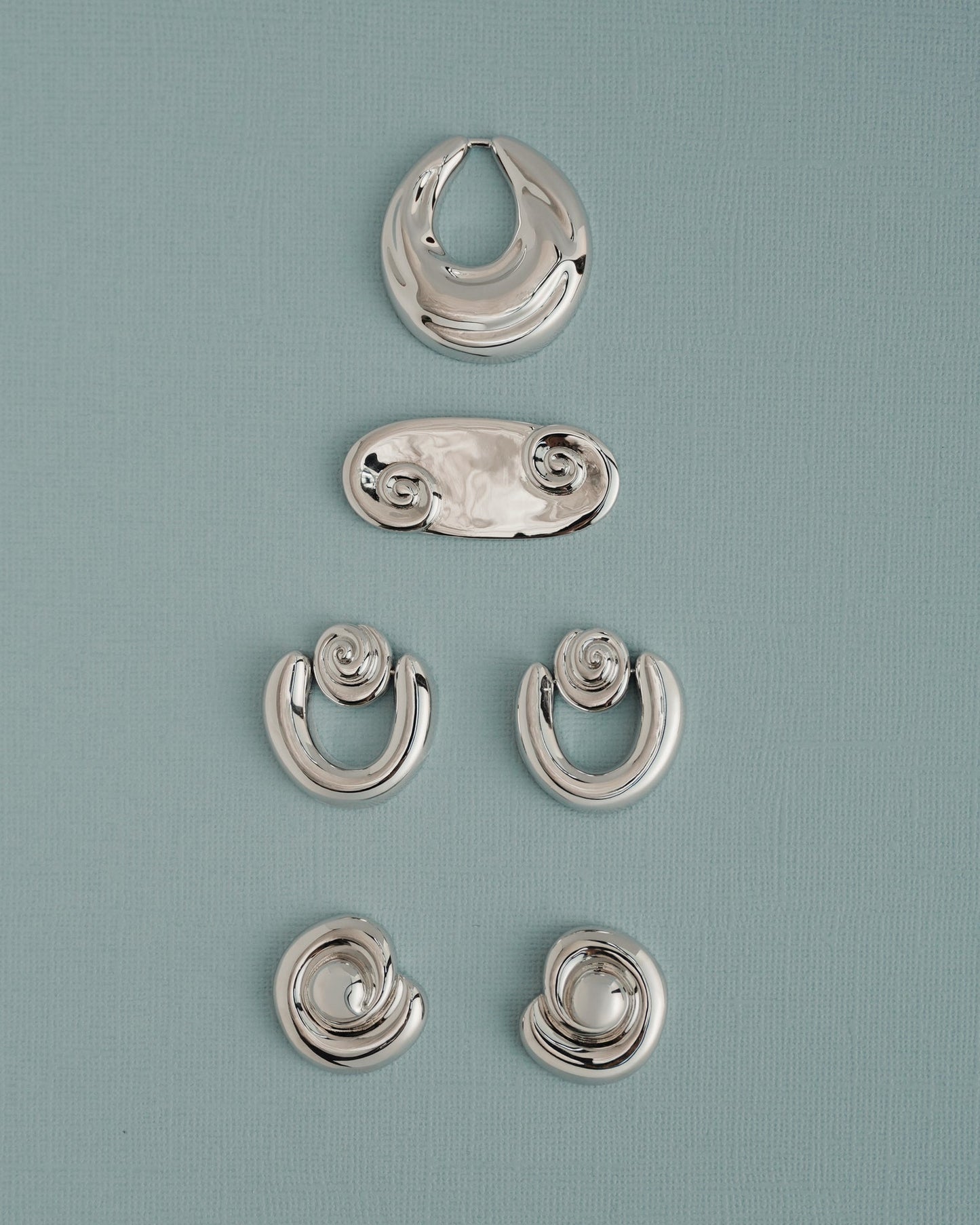 Five pairs of Sabine Statement Earrings by Mod + Jo, featuring shiny silver in abstract and spiral shapes, are displayed vertically on a light blue textured background.
