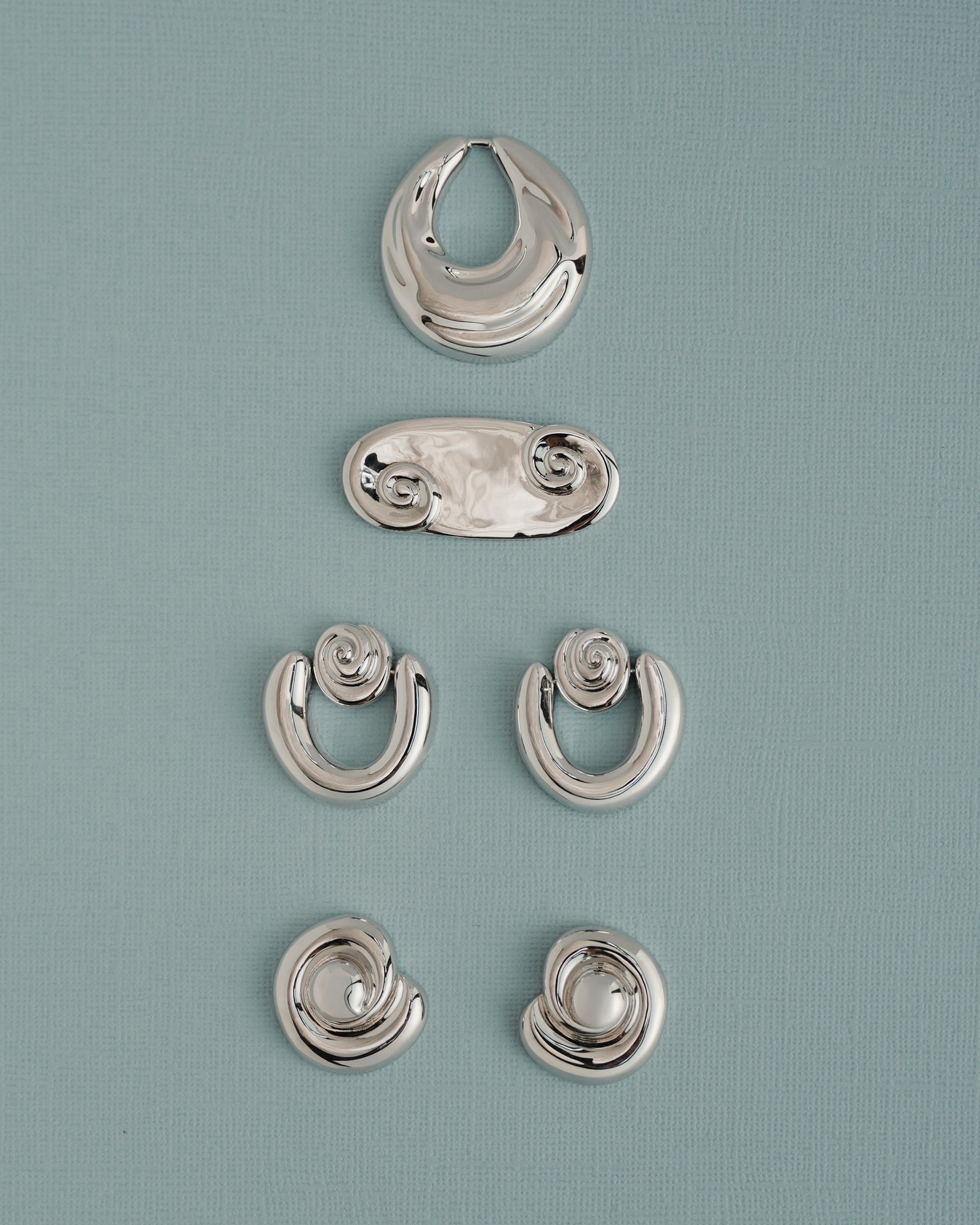 Five pairs of Sabine Statement Earrings by Mod + Jo, featuring shiny silver in abstract and spiral shapes, are displayed vertically on a light blue textured background.