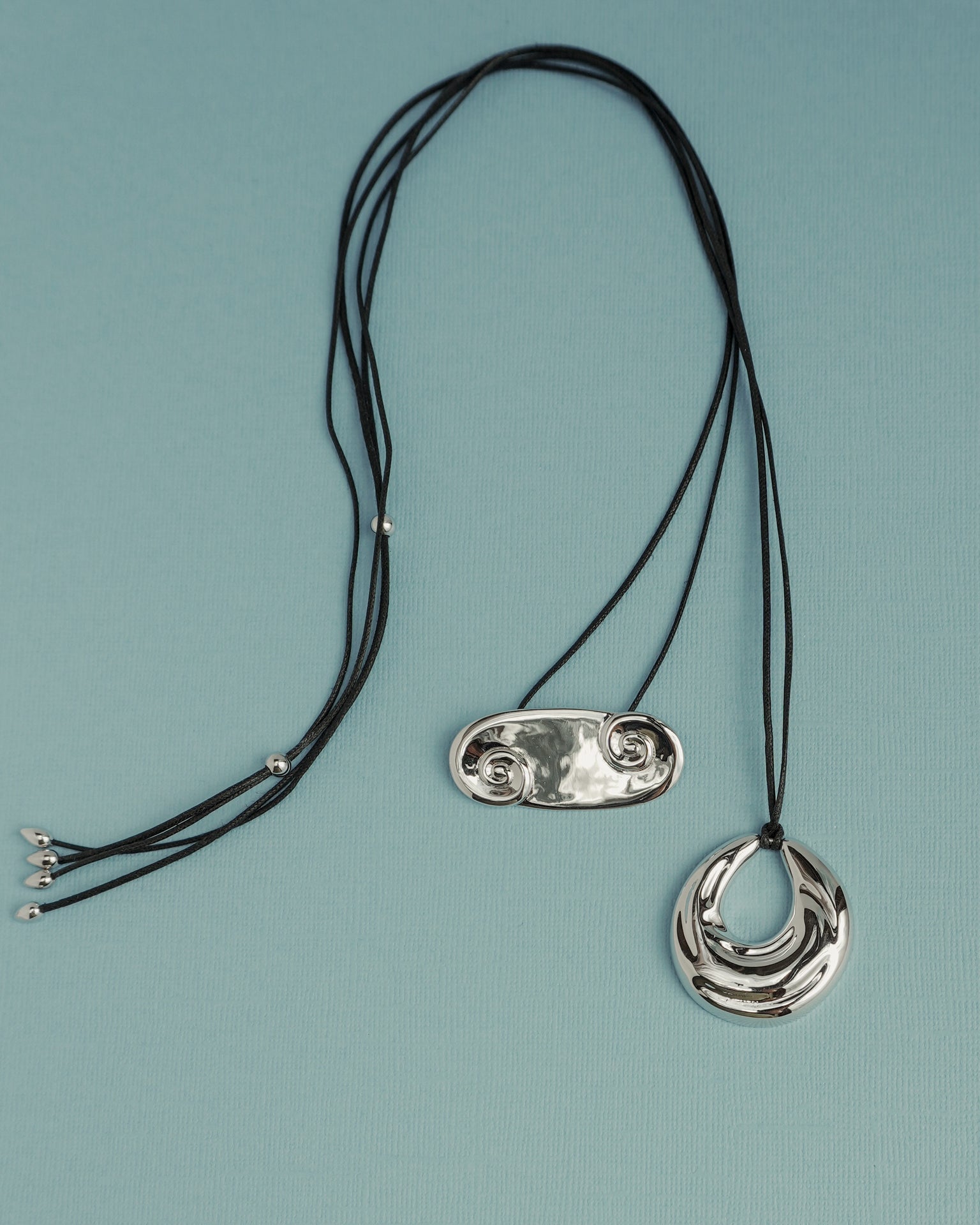 Two Suna Spiral Cord Necklaces by Mod + Jo rest on a light blue surface: one has a shiny double-spiral pendant, the other features a reflective crescent charm. Each adjustable piece blends unique style with comfort.