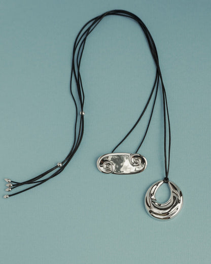 Two Suna Spiral Cord Necklaces by Mod + Jo rest on a light blue surface: one has a shiny double-spiral pendant, the other features a reflective crescent charm. Each adjustable piece blends unique style with comfort.