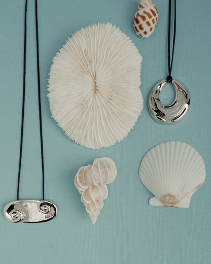 Mod + Jo’s Kira Cord Necklace is featured on a light blue background with white seashells—a round coral, scallop, and spiral shells—for a charming coastal-inspired display.