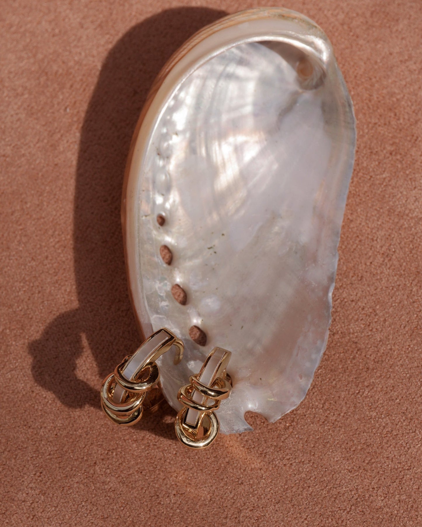A shiny mother of pearl seashell sits on sand, with a pair of Mod + Jo Levi Huggies—tarnish-resistant gold earrings—resting at its base.