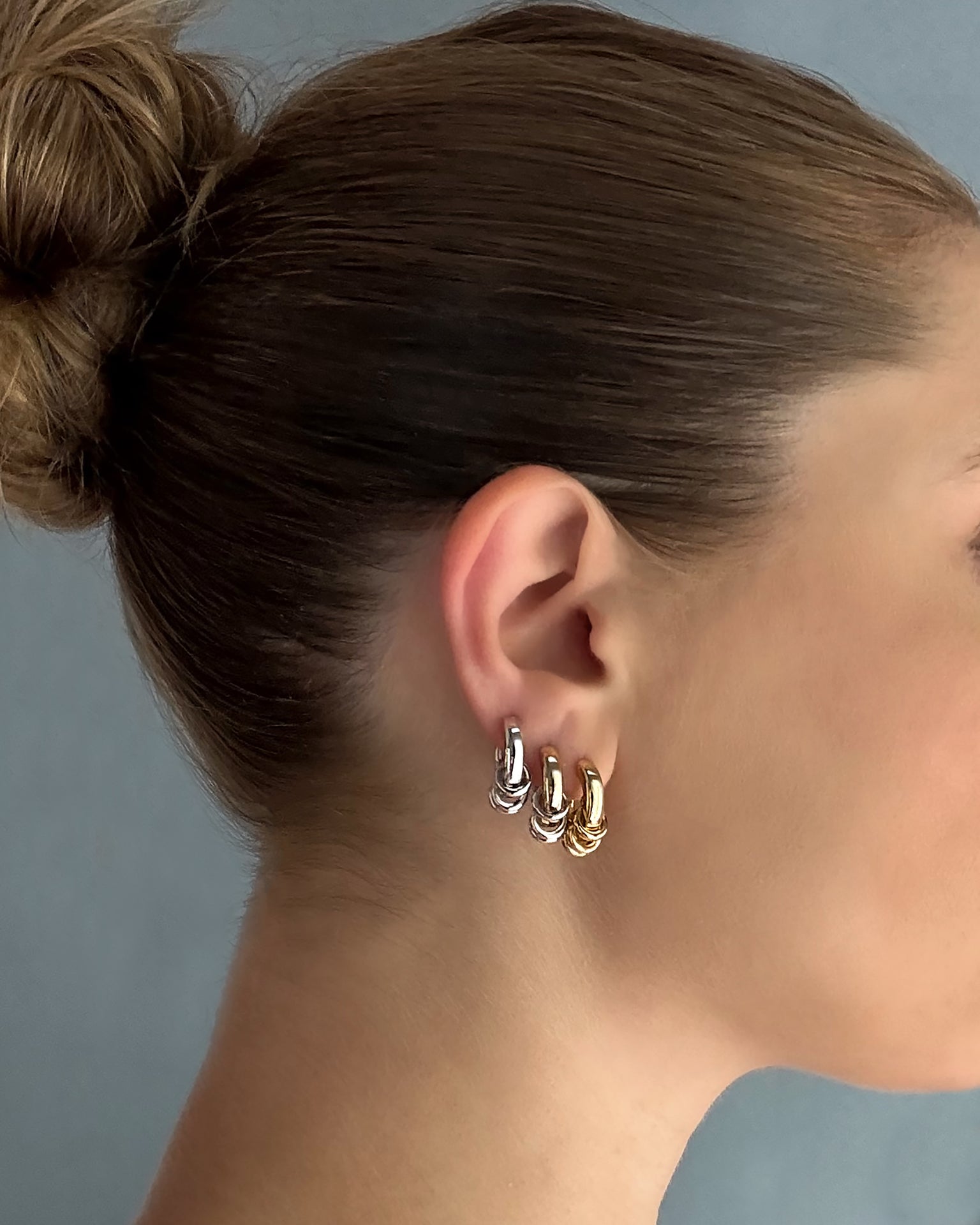 A woman with brown hair in a bun wears three small hoop earrings—including Mod + Jo’s Grande Levi Huggies in gold—on her left ear, alongside silver and rose gold hoops. She is facing right against a plain, soft blue background.