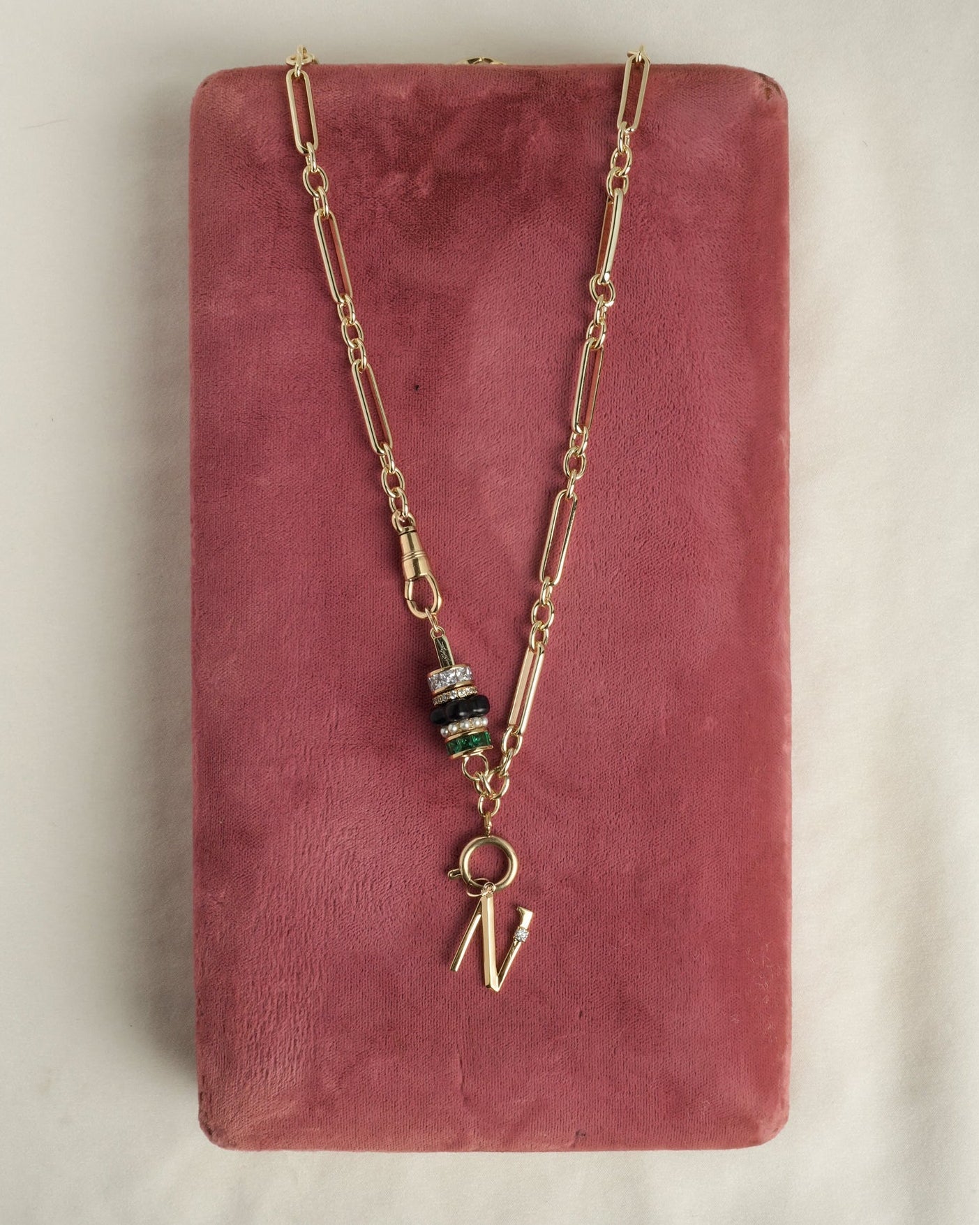 The Mod + Jo Liam Space Bar Chain Necklace features 14k gold plating, a cluster of black, clear, and green beads, and a gold letter N charm, displayed on a pink velvet rectangular box.