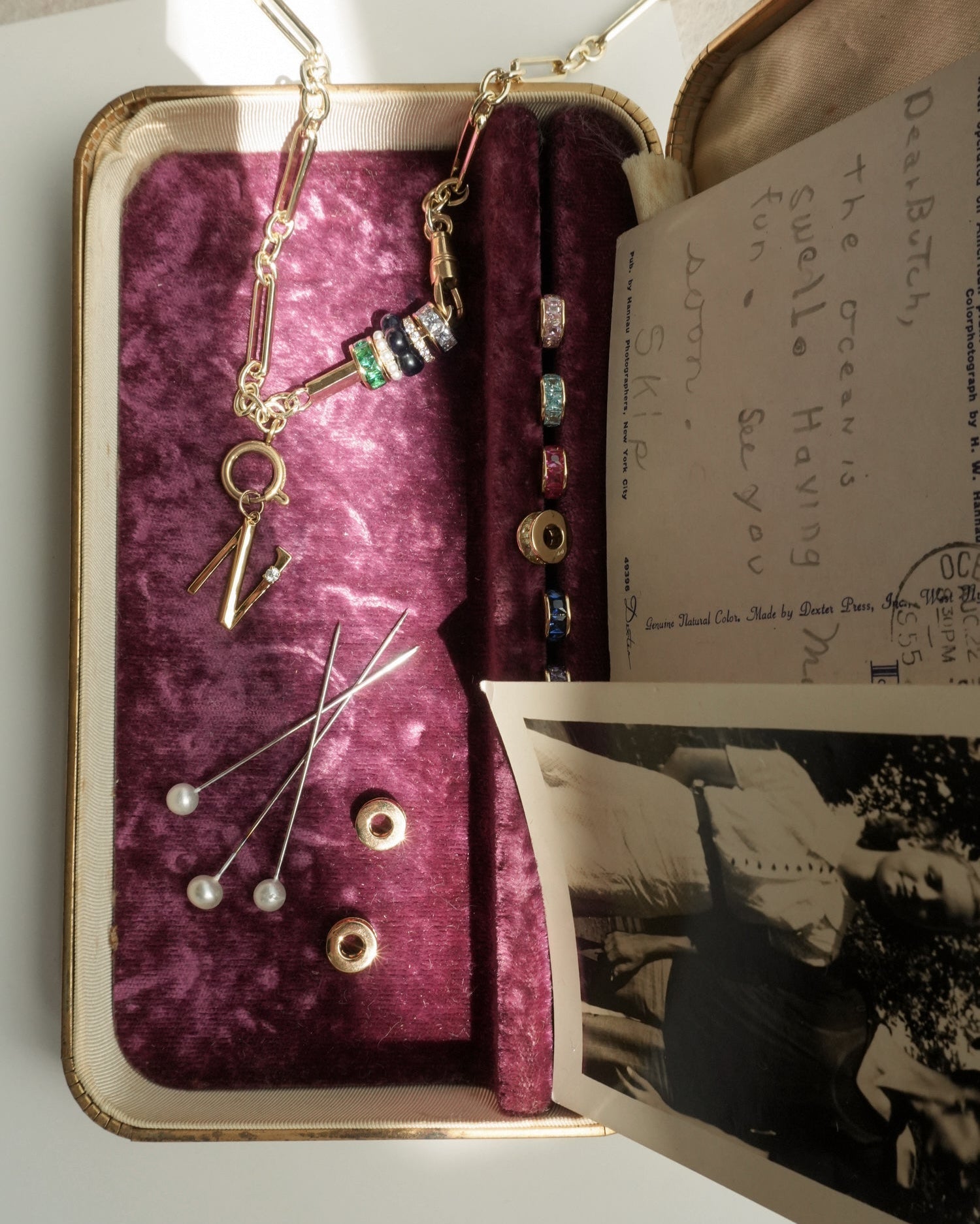 An open vintage jewelry box with velvet lining holds pins with pearl tops, colorful beads, the Mod + Jo Liam Space Bar Chain Necklace, an old black-and-white photo, and a handwritten postcard. Sunlight casts soft shadows.