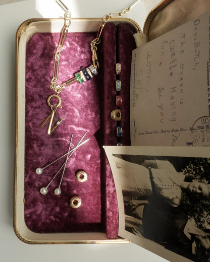 An open vintage jewelry box with velvet lining holds pins with pearl tops, colorful beads, the Mod + Jo Liam Space Bar Chain Necklace, an old black-and-white photo, and a handwritten postcard. Sunlight casts soft shadows.