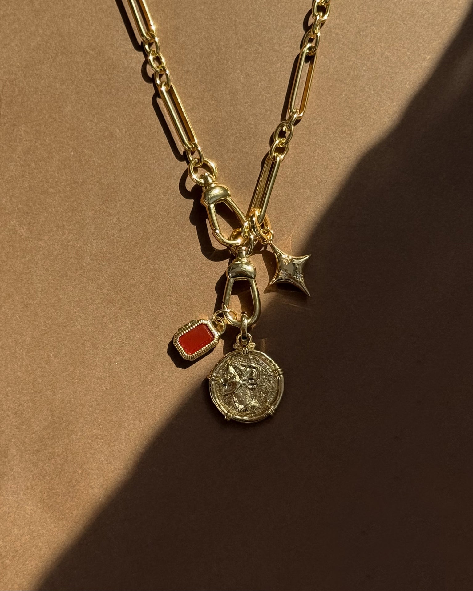 The Mod + Jo Naomi Ancient Coin Charm gold chain necklace features three unique charms—a coin, a red rectangular stone, and a small gold star—channeling Greek goddess style against a warm brown background with soft shadows.