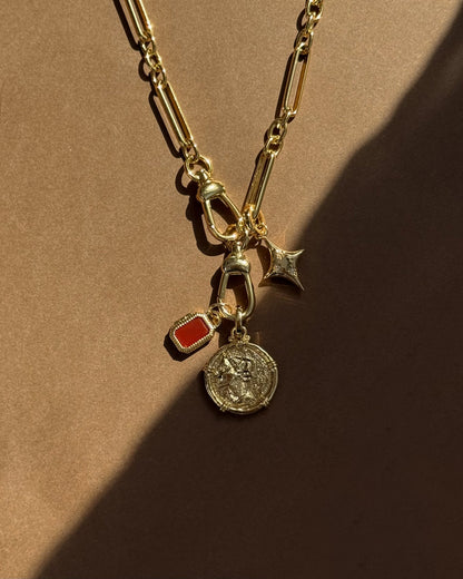 The Mod + Jo Naomi Ancient Coin Charm gold chain necklace features three unique charms—a coin, a red rectangular stone, and a small gold star—channeling Greek goddess style against a warm brown background with soft shadows.