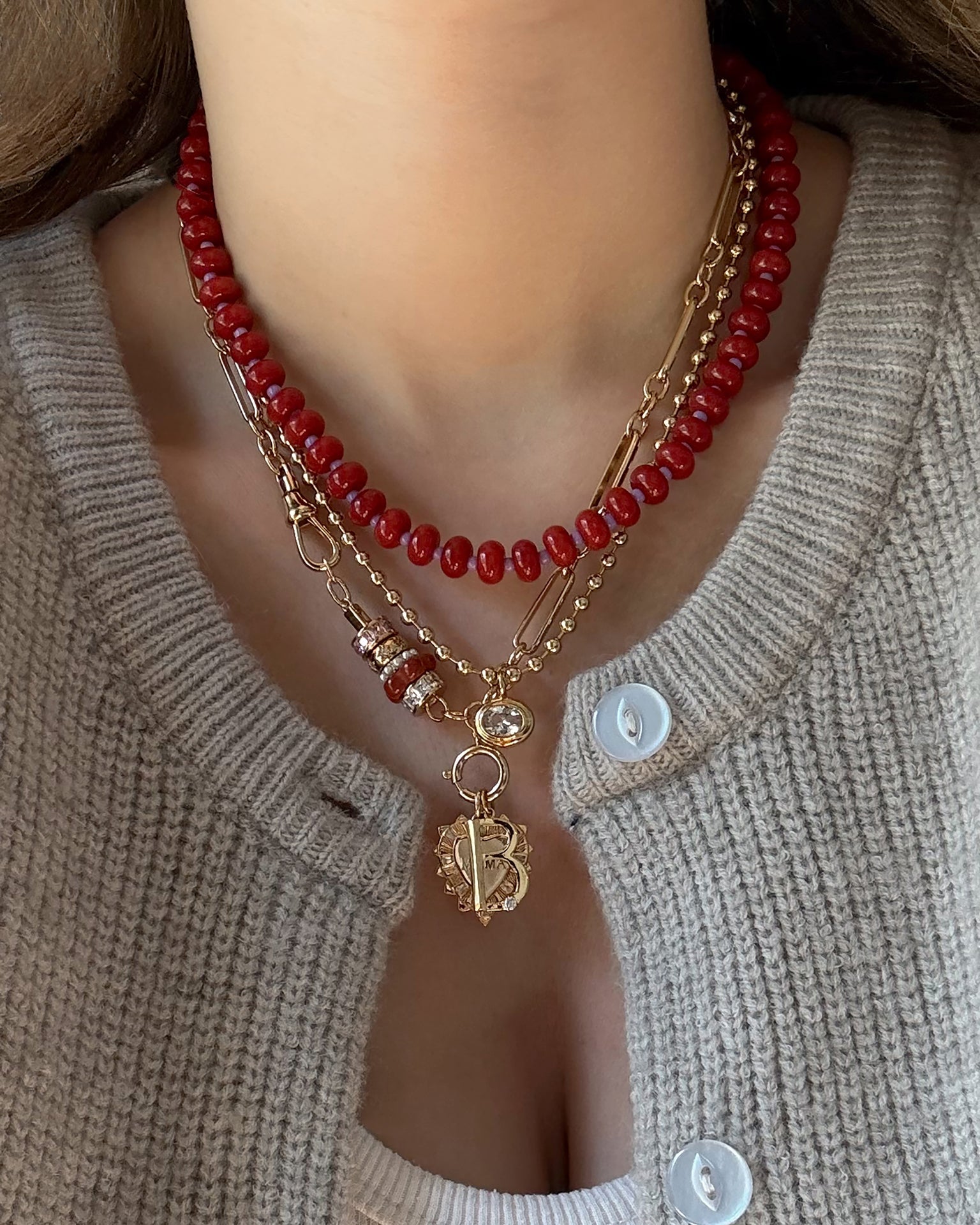 A person in a beige knit cardigan and white top layers necklaces, including the Mod + Jo Liam Space Bar Chain Necklace, a red beaded choker, gold chains, and pendants—one with the letter B.