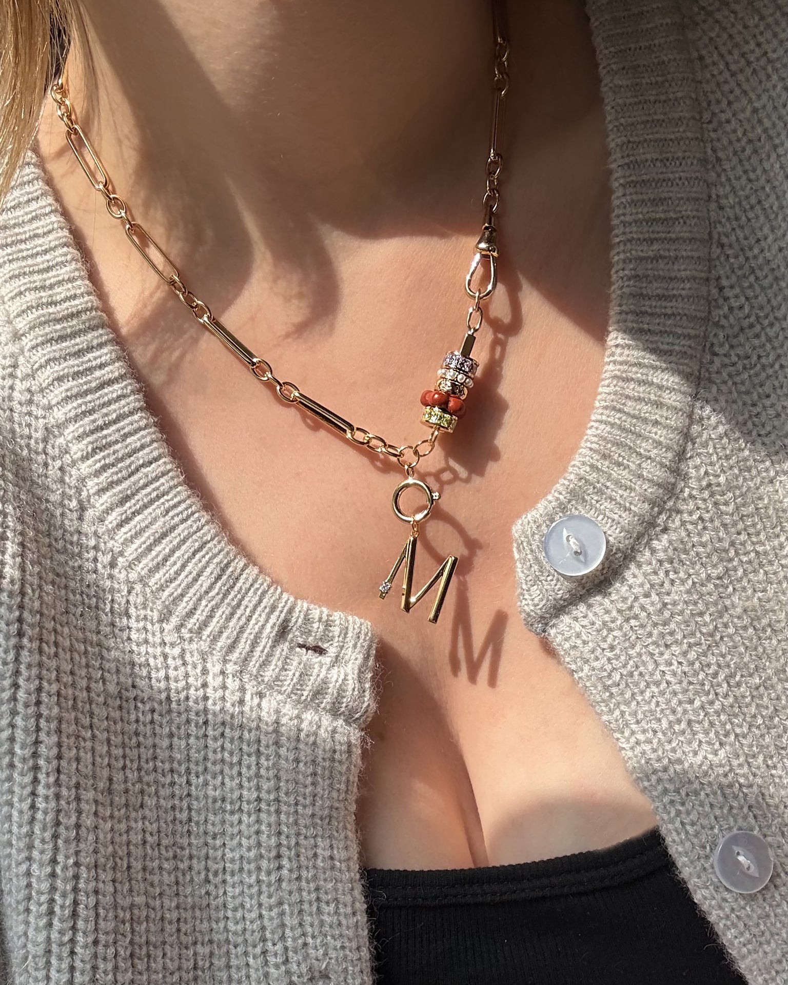 A person wearing a beige knit cardigan and the Mod + Jo Liam Space Bar Chain Necklace with colorful beads and a large gold letter M pendant. Sunlight casts the necklace's shadow onto their chest.