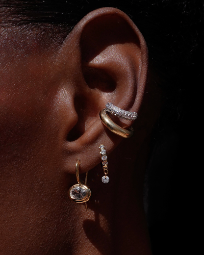 A close-up of an ear with four earrings: a gold hoop with CZ accents, the Nyla Pave Ear Cuff, a gold hoop with three dangling diamonds, and a gold earring featuring a large oval gemstone against a black background.