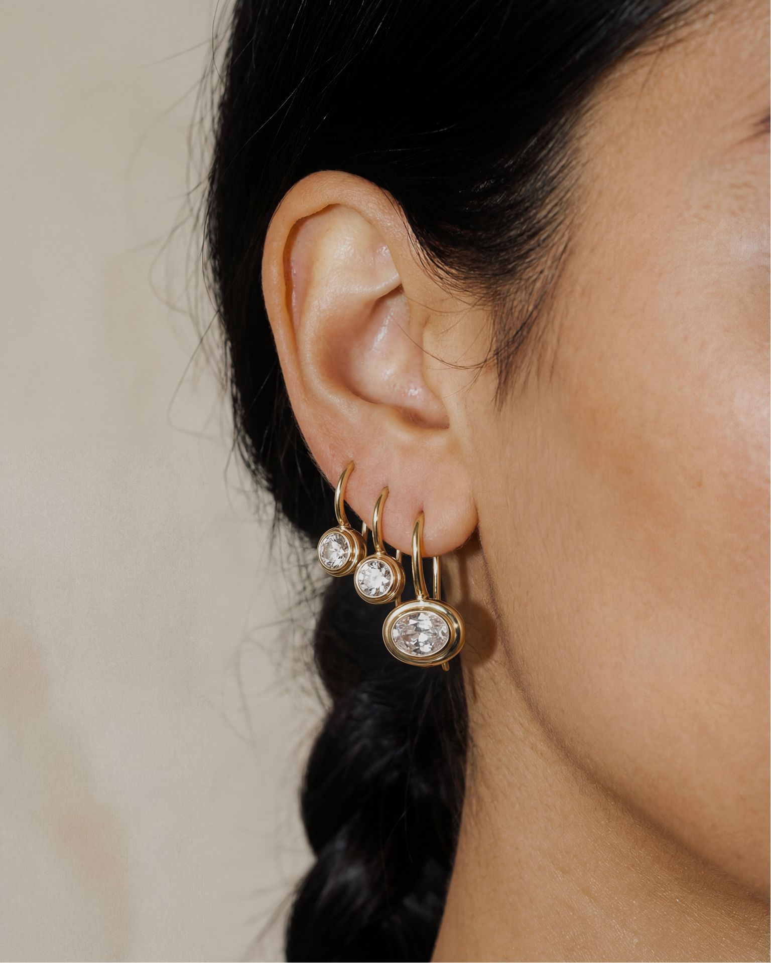 Close-up of an ear wearing gold hoop earrings with clear gemstones on a neutral background