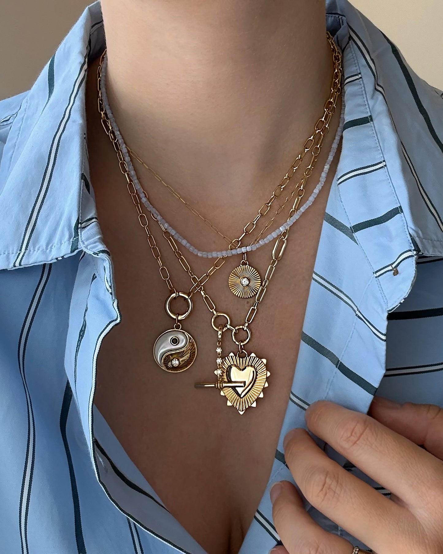 A person wearing a blue striped shirt and layered Mod + Jo gold necklaces, including the Grande Ava Yin Yang Charm, a sunburst pendant, a mother-of-pearl necklace, and a heart-shaped locket—one hand resting gently on their chest.