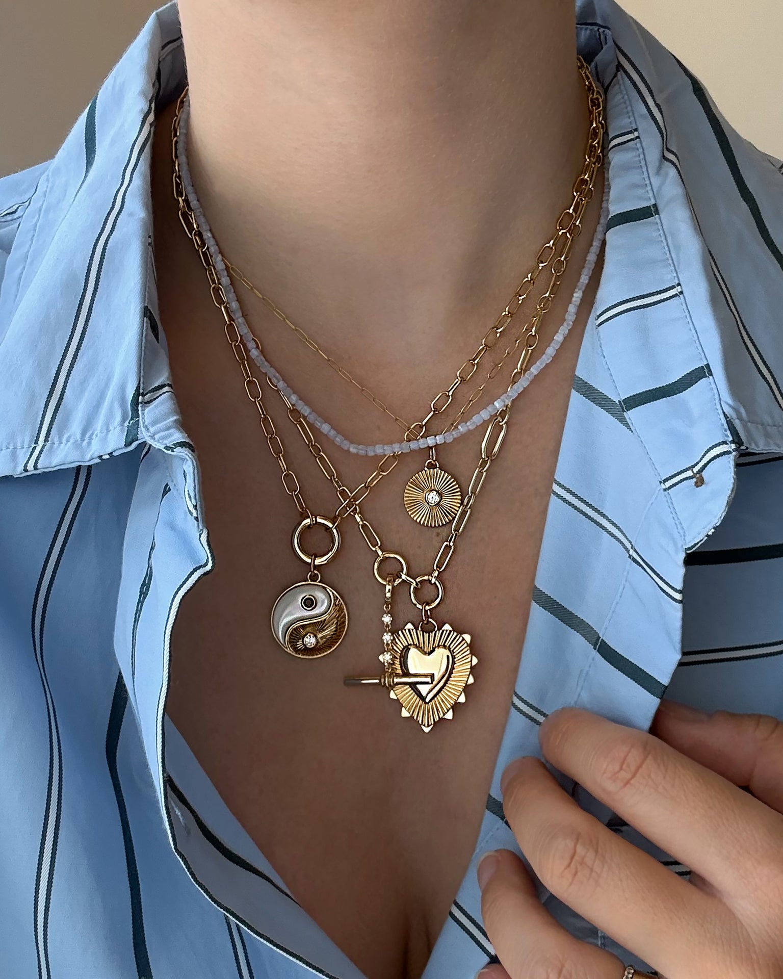 A person wearing a blue striped shirt and layered Mod + Jo gold necklaces, including the Grande Ava Yin Yang Charm, a sunburst pendant, a mother-of-pearl necklace, and a heart-shaped locket—one hand resting gently on their chest.