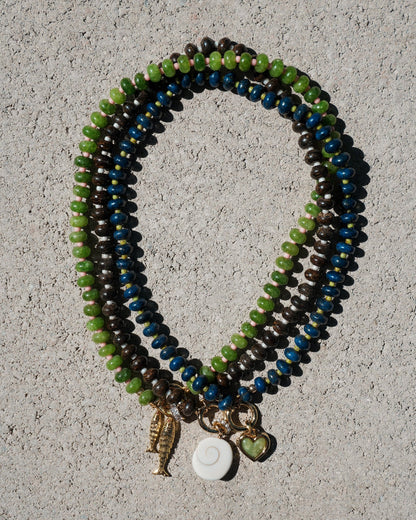 The Macey Gemstone Necklace by Mod + Jo rests on a textured gray surface, displaying green, blue, and brown beads—some with genuine gemstones like lapis lazuli—and gold charms: a fish, a white spiral, and a heart.