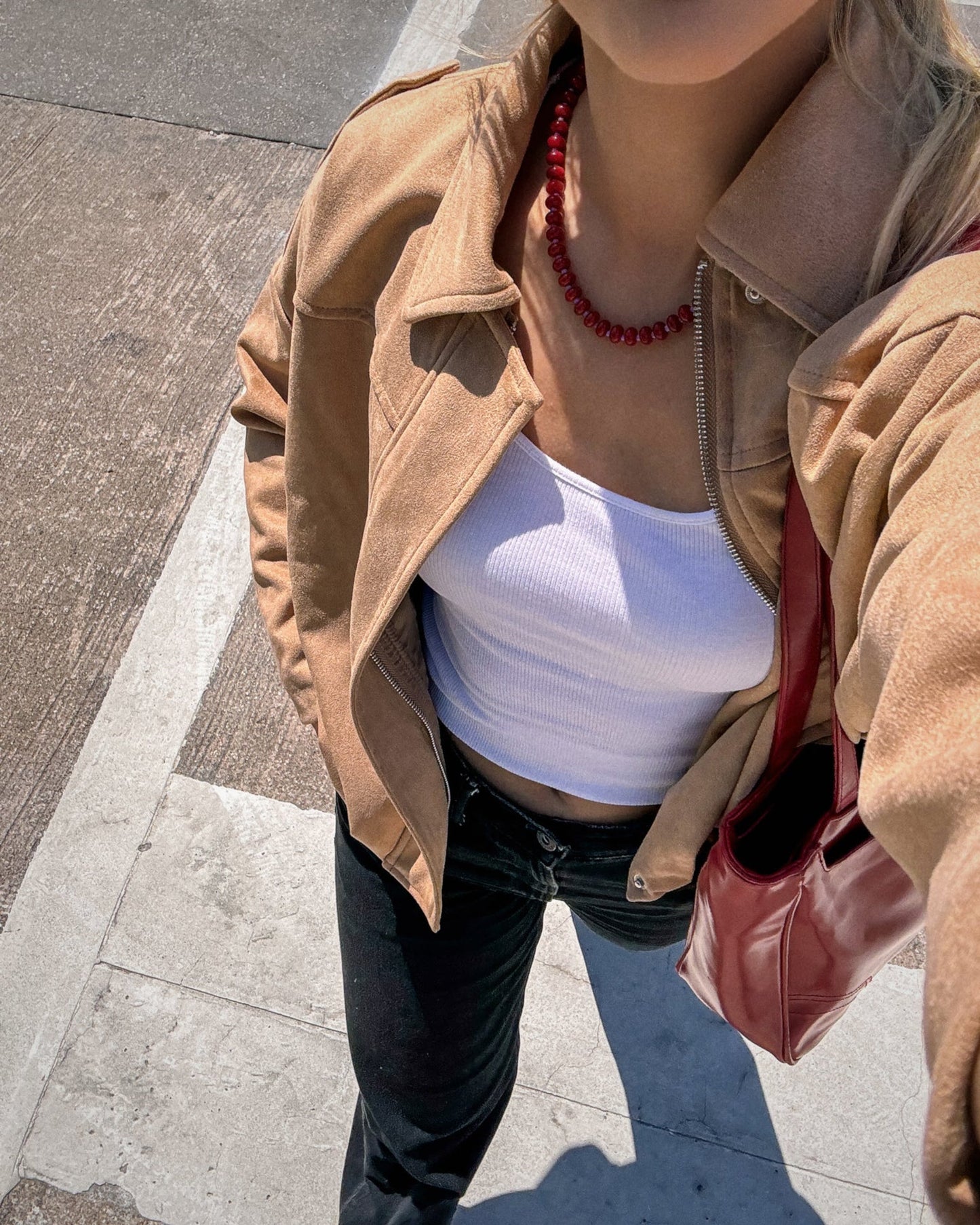 A woman stands on a sidewalk in a tan jacket, white ribbed crop top, black pants, and a red handbag. She accessorizes with the Mod + Jo Macey Gemstone Necklace. The photo is shot from above, showing her face and upper body.