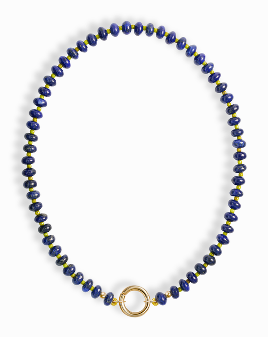 The Mod + Jo Macey Gemstone Necklace features round blue and gold-flecked lapis lazuli beads in a single strand, finished with a statement circular gold clasp at the center—all set against a white background.