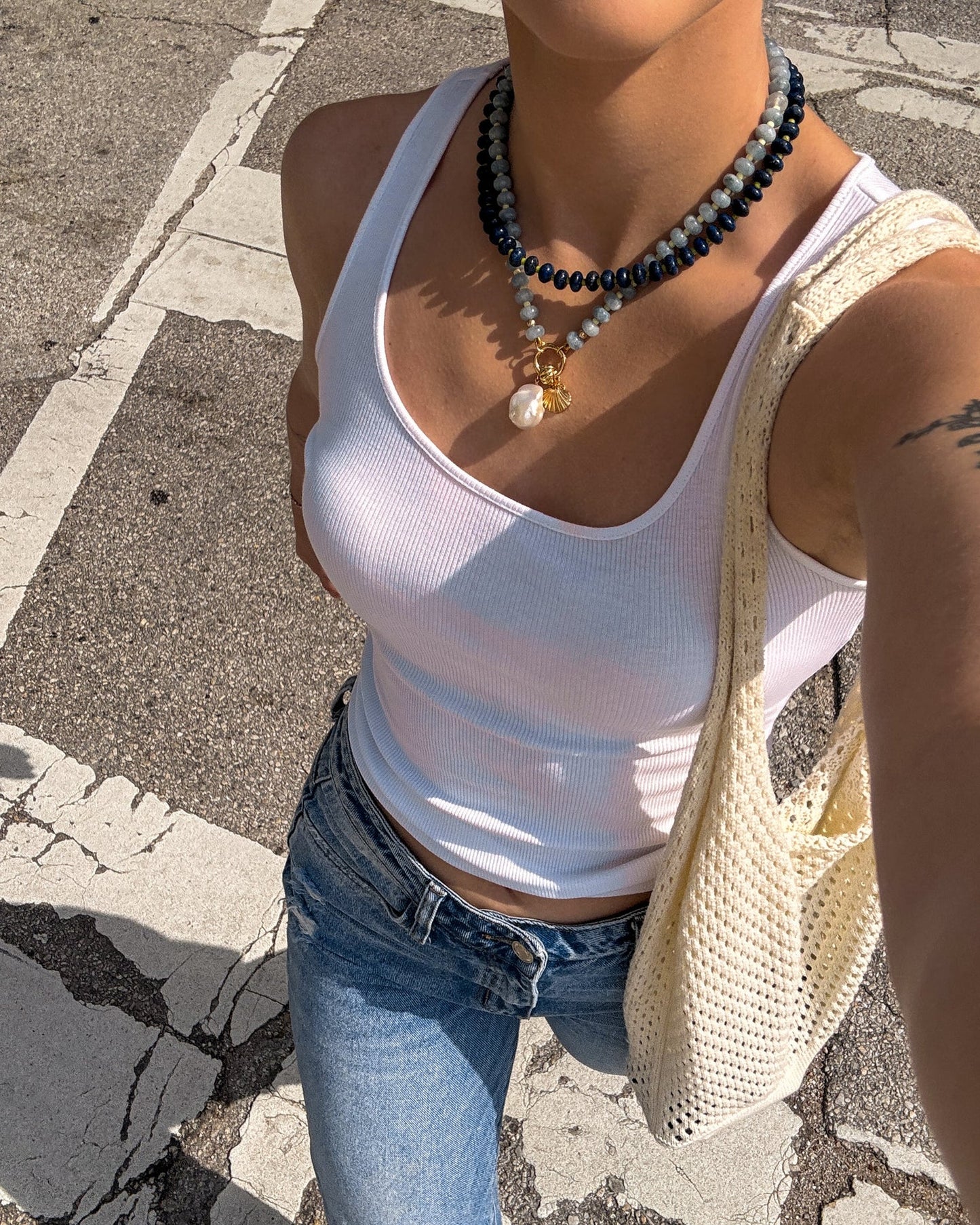 Wearing a white ribbed tank top, blue jeans, and a crochet shoulder bag, a person stands on a sunlit crosswalk. They accessorize with the Mod + Jo Macey Gemstone Necklace and display a partial arm tattoo.