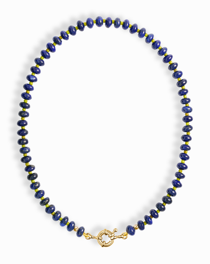 The Mod + Jo Macey Gemstone Necklace features round, deep blue lapis lazuli beads separated by small gold accents and finished with a circular gold clasp, arranged in an oval shape against a white background.