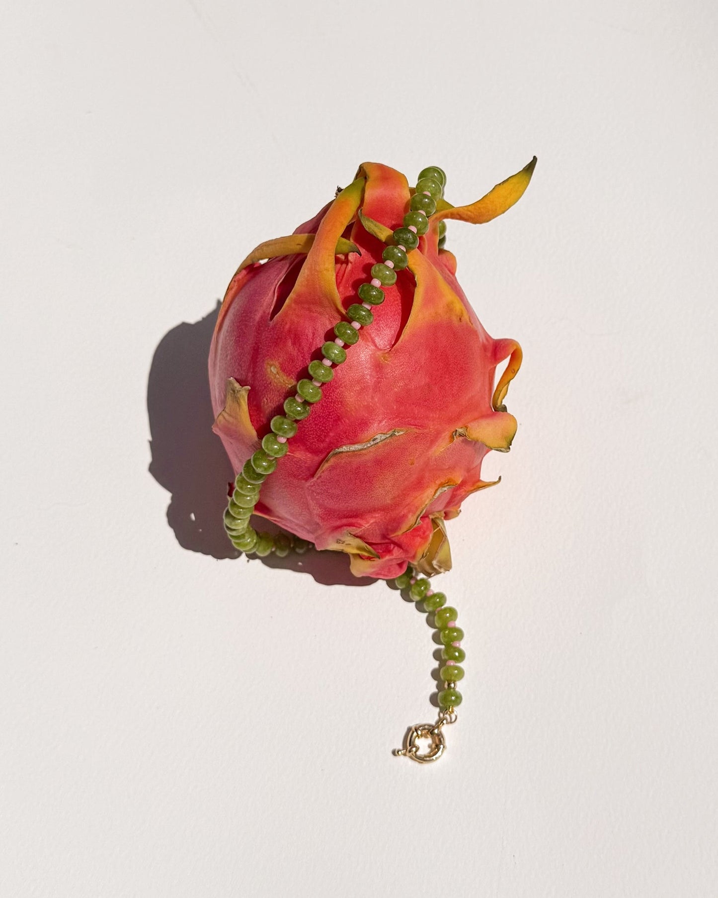 The Mod + Jo Macey Gemstone Necklace, featuring green jade and a gold clasp, is draped over a pink dragon fruit against a plain white background with a soft shadow.