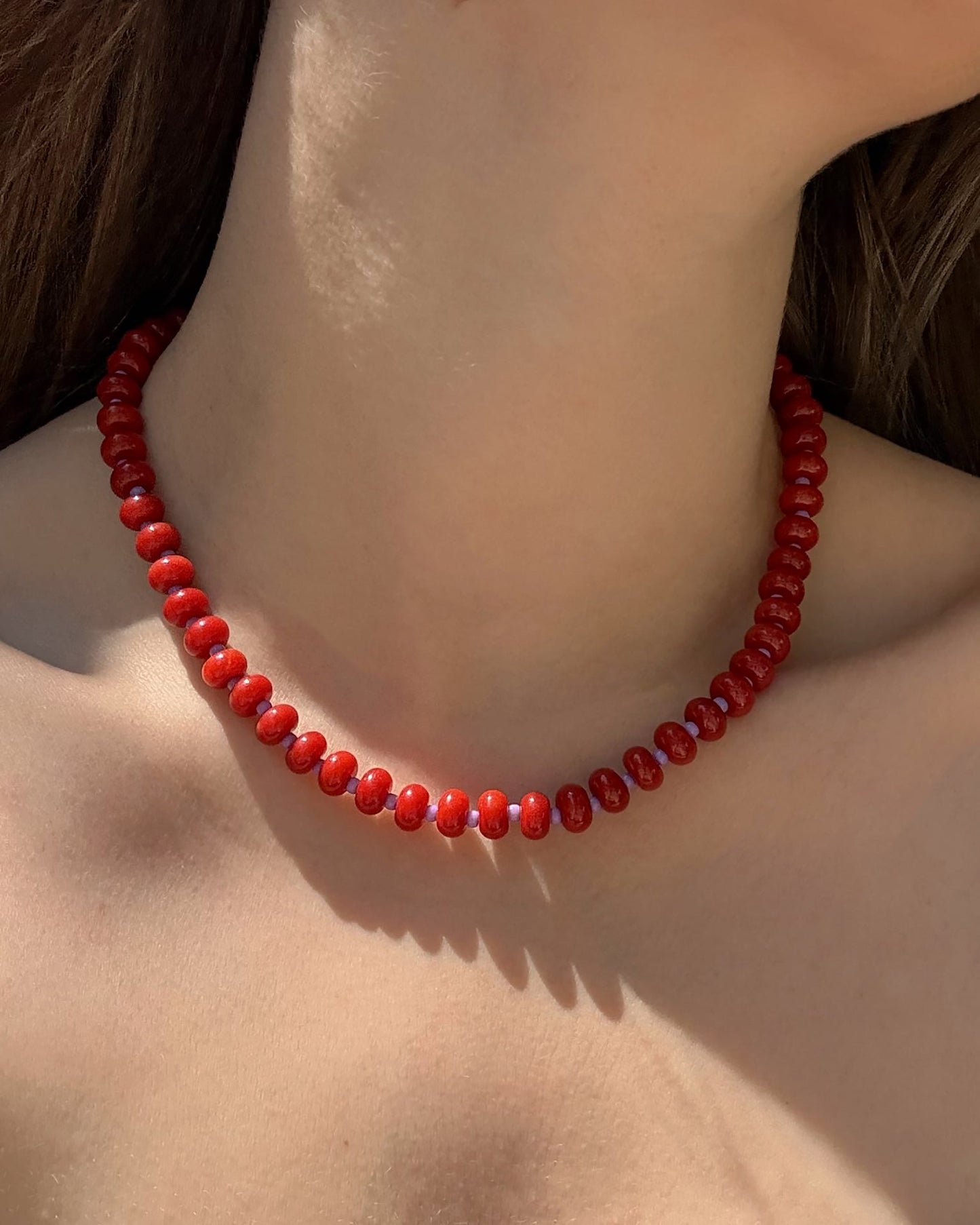 A close-up of a neck adorned with the Mod + Jo Macey Gemstone Necklace—a short, single-strand accessory featuring shiny red beads—sunlight illuminates the skin, with brown hair partly visible.