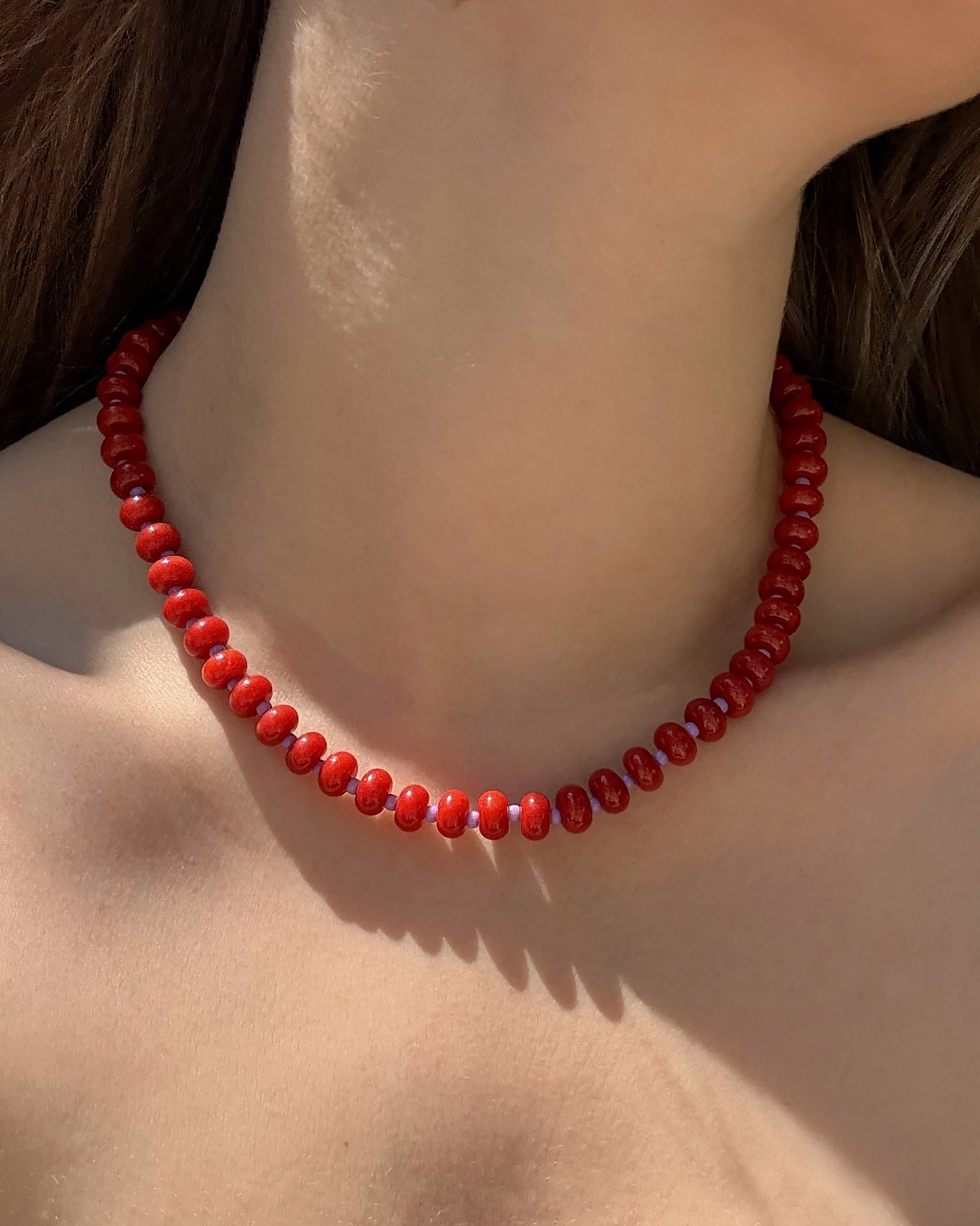 A close-up of a neck adorned with the Mod + Jo Macey Gemstone Necklace—a short, single-strand accessory featuring shiny red beads—sunlight illuminates the skin, with brown hair partly visible.