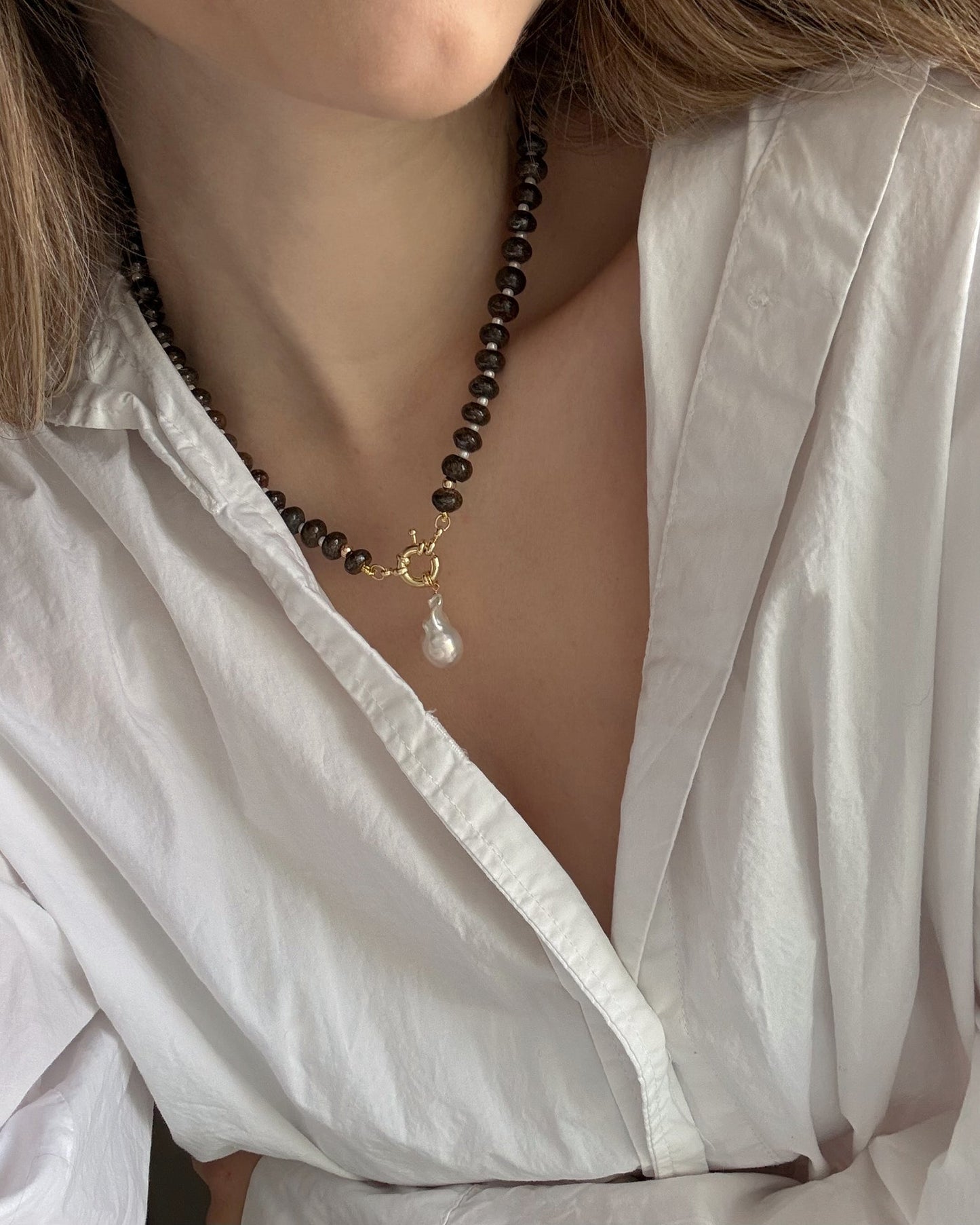 A person wears a white, open-collared blouse and the Mod + Jo Macey Gemstone Necklace, featuring genuine gemstones, a gold clasp, and a dangling pearl pendant for an elegant look.