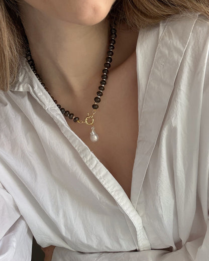 A person wears a white, open-collared blouse and the Mod + Jo Macey Gemstone Necklace, featuring genuine gemstones, a gold clasp, and a dangling pearl pendant for an elegant look.