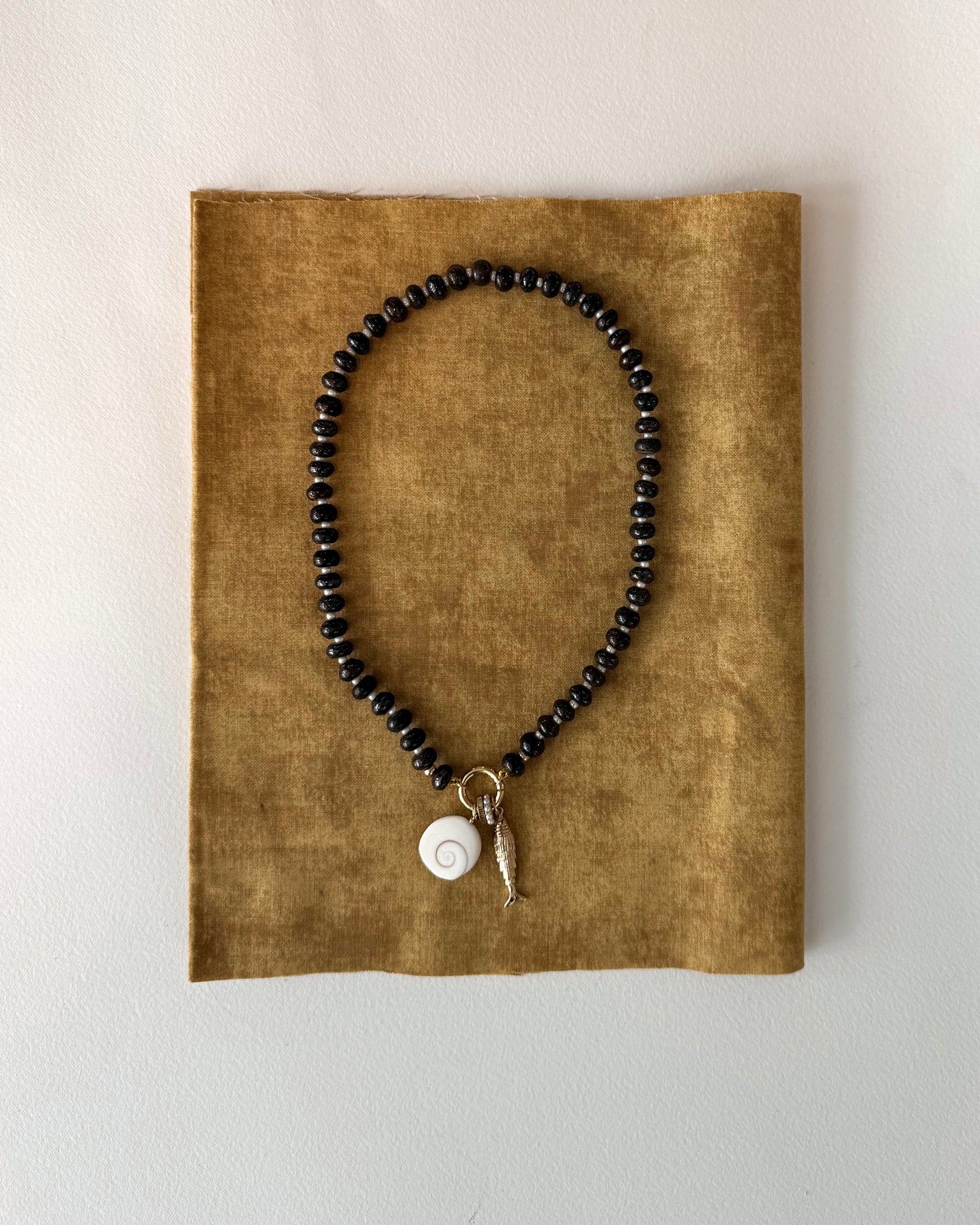 The Macey Gemstone Necklace by Mod + Jo features black beads, a white spiral shell pendant, and a small gold fish charm with 14k plating, showcased on textured brown fabric against a light background.