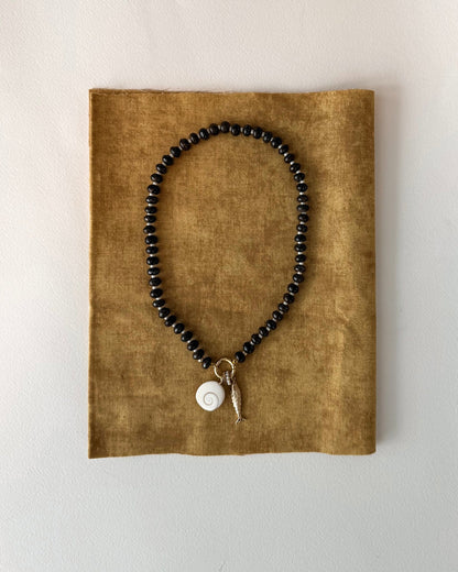 The Macey Gemstone Necklace by Mod + Jo features black beads, a white spiral shell pendant, and a small gold fish charm with 14k plating, showcased on textured brown fabric against a light background.
