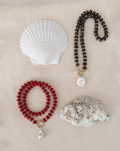 The Mod + Jo Macey Gemstone Necklace is shown on beige fabric, featuring black beads with a spiral shell pendant and red semi-precious stones with a pearl pendant, displayed near scallop and rough white shells.