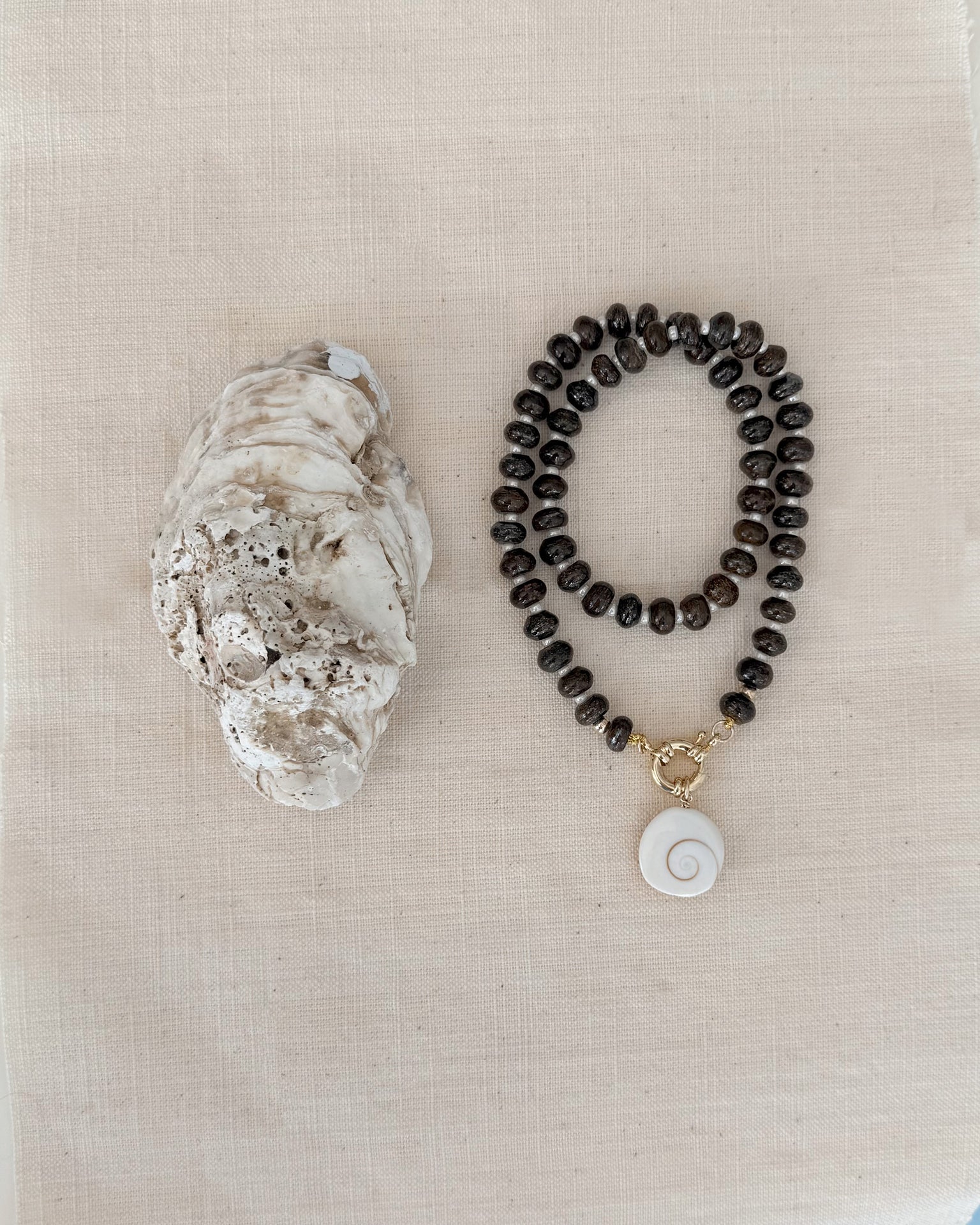 The Mod + Jo Macey Gemstone Necklace, featuring dark round beads, a white spiral shell pendant, and genuine gemstones, is displayed beside a large oyster shell on a light beige fabric background.