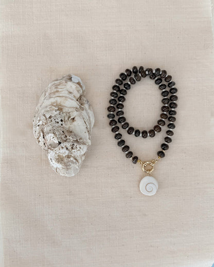 The Mod + Jo Macey Gemstone Necklace, featuring dark round beads, a white spiral shell pendant, and genuine gemstones, is displayed beside a large oyster shell on a light beige fabric background.