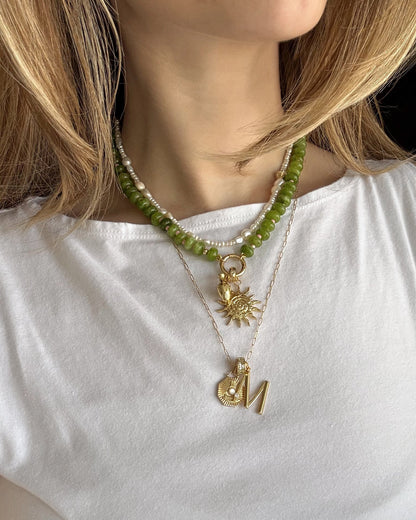 A woman with shoulder-length blonde hair wears a white shirt and the Mod + Jo Macey Gemstone Necklace—a layered set featuring a green jade beaded choker, a gold sun pendant, and a gold “M” with decorative charm.